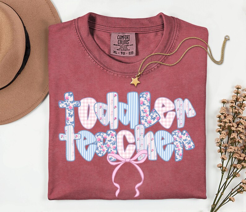 May include: A maroon Comfort Colors t-shirt with the words "Toddler Teacher" in a floral, gingham, and striped design. A gold star pendant necklace and a tan hat are also visible.