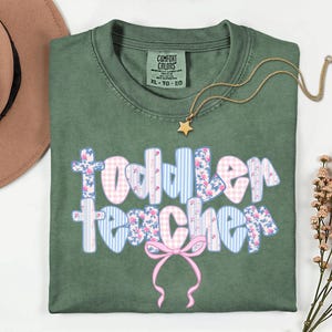 May include: An olive green Comfort Colors t-shirt with the words "Toddler Teacher" in a floral and gingham design. A gold star pendant necklace and a brown hat are also in the image.