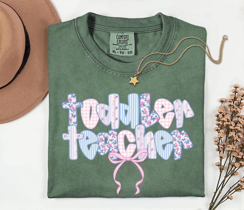 May include: An olive green Comfort Colors t-shirt with the words "Toddler Teacher" in a floral and gingham design. A gold star pendant necklace and a brown hat are also in the image.