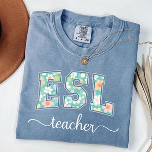 May include: A blue Comfort Colors t-shirt with the text "ESL teacher" in white script. The letters "ESL" are decorated with a floral pattern in shades of pink, green, and white. A gold necklace with a pendant is draped across the shirt.