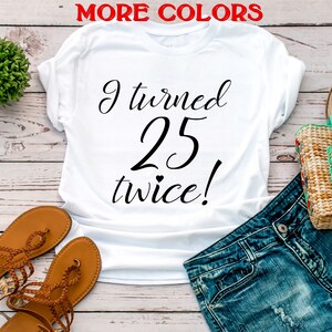 May include: White t-shirt with black text that reads "I turned 25 twice!" The shirt is laid out on a wooden surface with a pair of brown sandals and blue jeans.