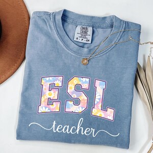 May include: A blue t-shirt with the letters "ESL" in a floral design, with the word "teacher" in a cursive font below. A gold necklace with a pendant is on the shirt. The shirt is folded on a white surface.