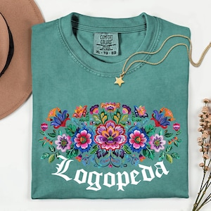 May include: Teal Comfort Colors t-shirt with a floral design in vibrant colors and the word "Logopeda" in white script. A gold star pendant necklace rests on the shirt. A brown hat is visible in the upper left corner.