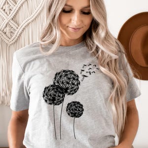 May include: A light gray t-shirt featuring a black dandelion design. The graphic shows three dandelions with seeds blowing away. The shirt is a crew neck and has short sleeves.