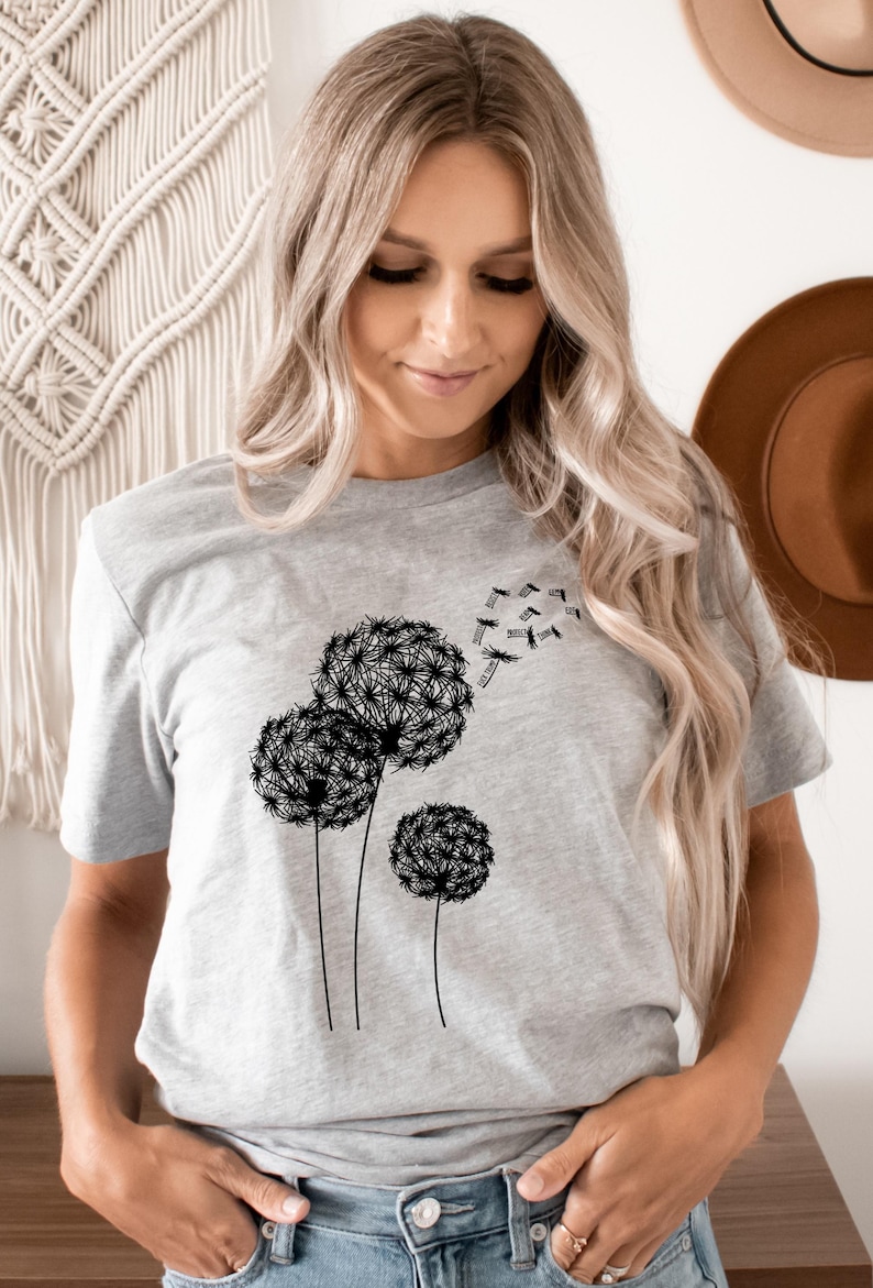May include: A light gray t-shirt featuring a black dandelion design. The graphic shows three dandelions with seeds blowing away. The shirt is a crew neck and has short sleeves.