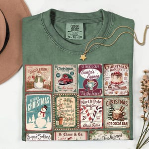 May include: Green Comfort Colors t-shirt with a Christmas-themed graphic. The graphic features vintage-style signs for Cand's Candy, Christmas Tree Farm, St. Nick's Cafe, and Christmas Bakery. A gold star pendant necklace is draped on the shirt.