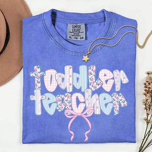 May include: A blue Comfort Colors t-shirt with the words "Toddler Teacher" in a floral, gingham, and striped design. A gold necklace with a star pendant rests on the shirt. A pink bow is at the bottom of the text.