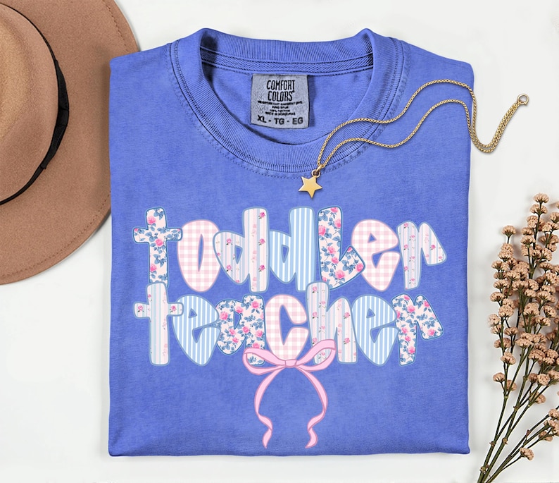 May include: A blue Comfort Colors t-shirt with the words "Toddler Teacher" in a floral, gingham, and striped design. A gold necklace with a star pendant rests on the shirt. A pink bow is at the bottom of the text.