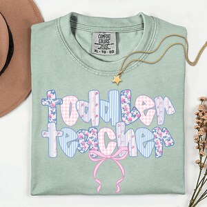 May include: A light green Comfort Colors t-shirt with the words "Toddler Teacher" in a floral and gingham design. A gold star necklace and a pink bow are also featured. The shirt is size XL-TG-EG.