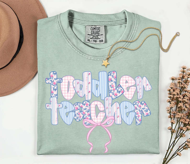 May include: A light green Comfort Colors t-shirt with the words "Toddler Teacher" in a floral and gingham design. A gold star necklace and a pink bow are also featured. The shirt is size XL-TG-EG.