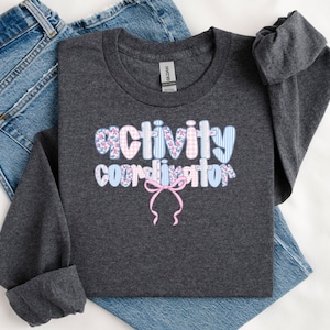 May include: A dark charcoal gray long-sleeve shirt featuring the text "activity coordinator" in a multi-patterned font with floral, gingham, and striped designs in shades of pink and blue, accented by a pink ribbon bow graphic below the text.