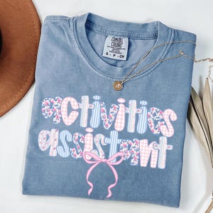 May include: A blue Comfort Colors t-shirt with the words "activities assistant" in a floral, gingham, and striped design. A pink bow is below the text. A gold necklace with a pendant is on the shirt.