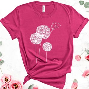 May include: A magenta t-shirt featuring a white dandelion design. The graphic shows three dandelion seed heads with seeds blowing away. The shirt has rolled-up sleeves and a knot at the bottom.