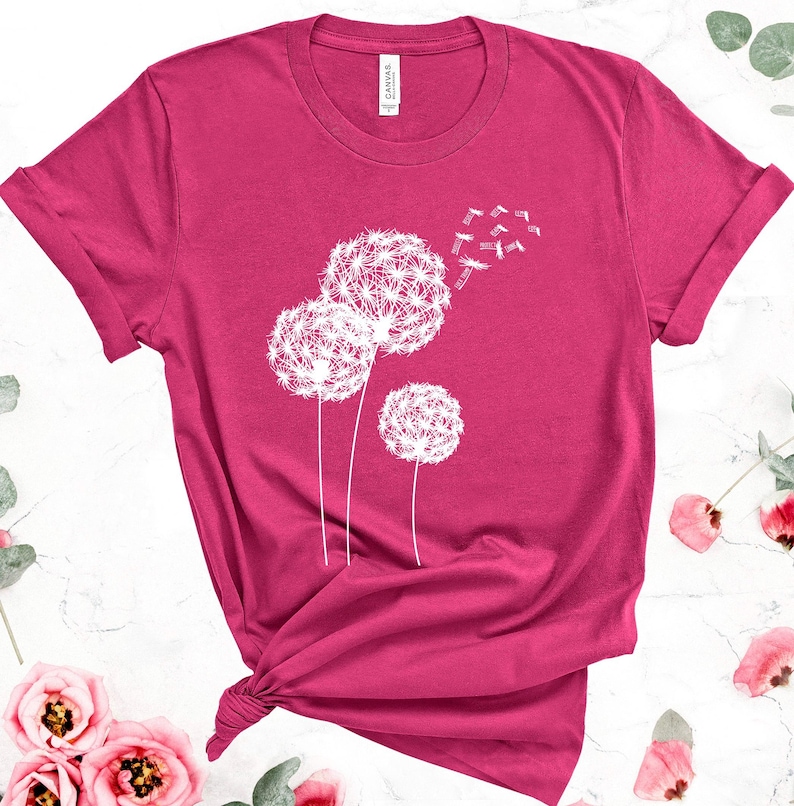 May include: A magenta t-shirt featuring a white dandelion design. The graphic shows three dandelion seed heads with seeds blowing away. The shirt has rolled-up sleeves and a knot at the bottom.