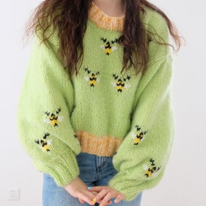 Può includere: Un maglione di lana verde chiaro con un bordo giallo e disegni di api ricamate.