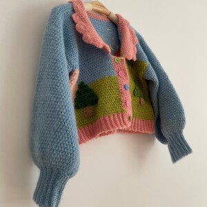 May include: A handmade, blue, pink, and green crochet cardigan with a scalloped collar. The sweater features a landscape design with trees and colorful buttons. The sleeves are a light blue color.