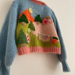 May include: A light blue crochet sweater with a landscape design. The design features a mountain, trees, a house, and a carrot. The sweater has a pink and light blue trim and long sleeves.