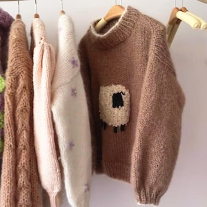 Op de afbeelding: Een verzameling gebreide truien die aan houten hangers hangen. De truien zijn in verschillende kleuren, waaronder een lichtbruine trui met een schapenontwerp, een lichtroze trui en een getextureerde bruine trui. De truien zijn gemaakt van zacht, gebreid materiaal.
