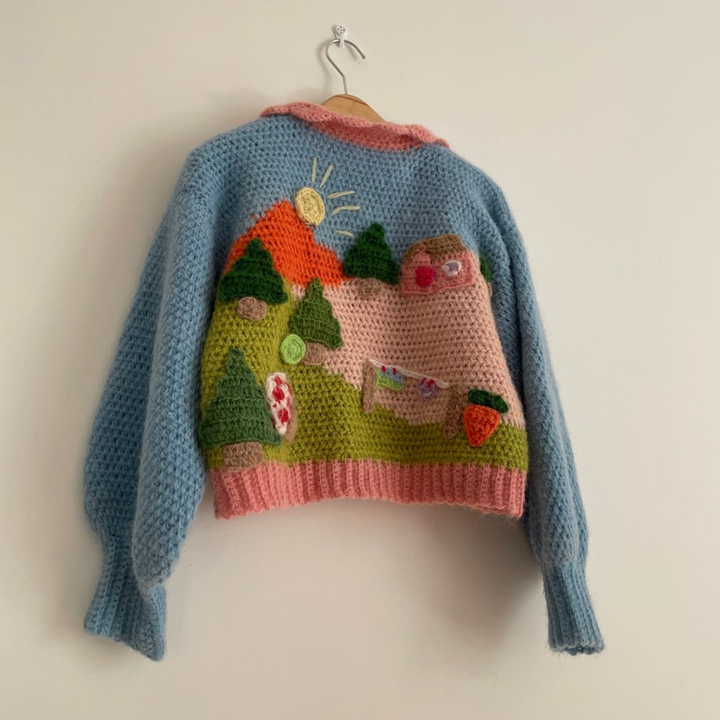 May include: A light blue crochet sweater with a landscape design on the back. The design includes a sun, mountains, trees, a house, and a carrot. The sweater has a pink collar and bottom hem.