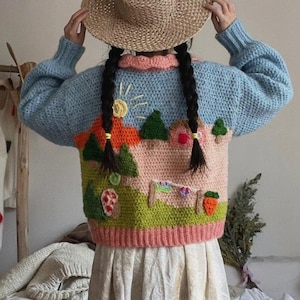 May include: A light blue crochet sweater with a landscape design. The sweater features a pink and orange house, green trees, and a sun. The sweater has a pink collar and cuffs. The person is wearing a straw hat.