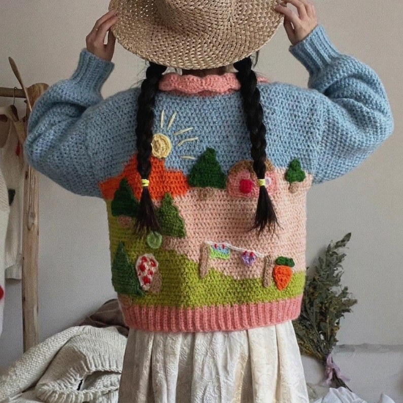 May include: A light blue crochet sweater with a landscape design. The sweater features a pink and orange house, green trees, and a sun. The sweater has a pink collar and cuffs. The person is wearing a straw hat.