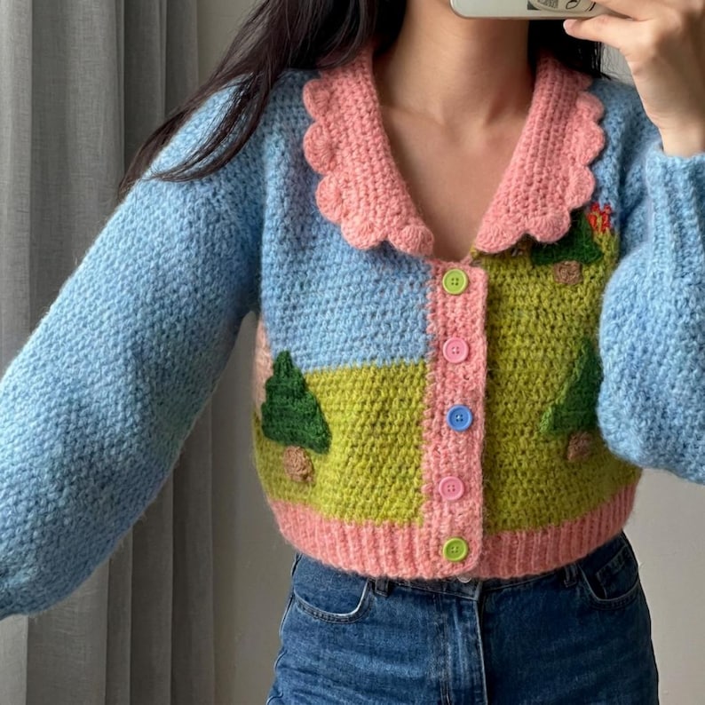 May include: A handmade, crochet cardigan in shades of blue, pink, and green. The sweater features a scalloped pink collar and a landscape design on the front, with button closures. The sleeves are a solid light blue.