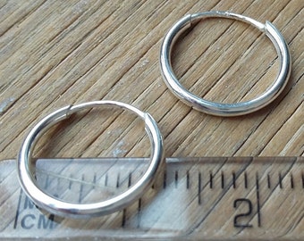 May include: Two silver hoop earrings, one is placed on top of a wooden ruler with centimeter markings. The ruler shows the earrings are about 1 cm in diameter.