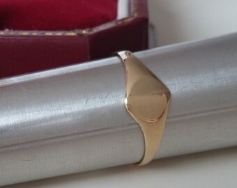 May include: A gold signet ring with an oval face is displayed on a silver metal rod. The ring is smooth and polished, reflecting light. A burgundy jewellery box with a gold clasp is in the background.