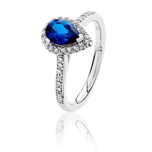 May include: A silver ring with a pear-cut, deep blue gemstone at its centre, encircled by a halo of smaller, clear stones. The ring's band is also embellished with small, clear stones. The ring is displayed against a white backdrop.