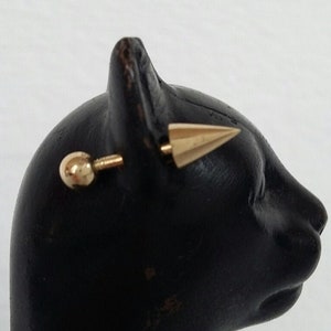 May include: Gold-toned ear piercing with a small ball on one end and a pointed tip on the other end. The piercing is displayed on a black mannequin head.