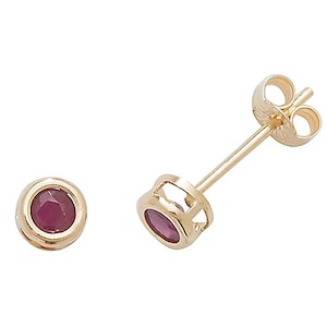 May include: A pair of gold stud earrings, each featuring a round, deep red gemstone set in a bezel. One earring is upright, showing the post and butterfly clasp. The other lies flat, displaying the gemstone.