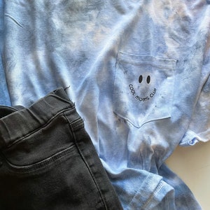 May include: A light blue tie-dye t-shirt with a pocket on the front. The pocket has a black smiley face with the text "Cool Moms Club" written around it.