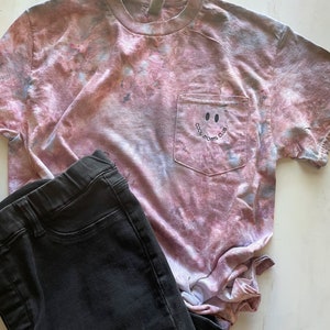 May include: A pink and grey tie-dye t-shirt with a pocket on the front. The pocket has a black smiley face and the text "Cool Moms Club".