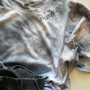 May include: A light blue tie-dye t-shirt with the word "mama" embroidered in black on the front. The shirt is folded and lying on a white surface.