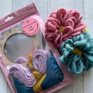 May include: A pink plastic bag containing a wooden flower loom, yarn in pink, yellow, and blue, and two finished flower crafts in pink and blue.