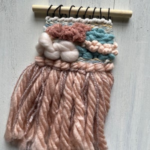 May include: A small, handmade woven wall hanging. The textile art piece features a light wooden dowel at the top with black loops for hanging. The woven design incorporates various textures and colors, including pink, cream, and blue yarns, creating a soft, tactile appearance.
