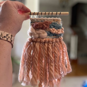May include: A small, handmade woven wall hanging. The textile art piece features a wooden dowel at the top with black thread accents. The woven design incorporates various textures and colors, including peach, blue, and cream-colored yarns, with a fringe of peach-colored yarn.
