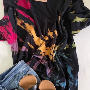 May include: A black tie-dye t-shirt with a colourful splatter pattern. The shirt is paired with blue jeans and black sandals.