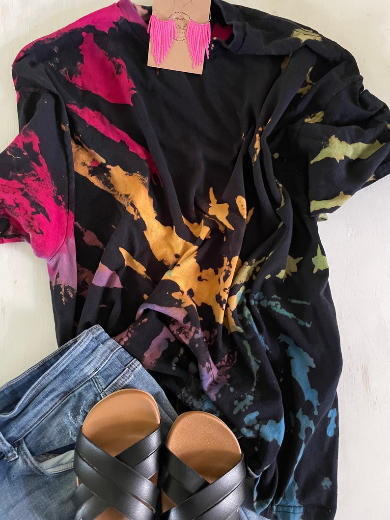 May include: A black tie-dye t-shirt with a colourful splatter pattern. The shirt is paired with blue jeans and black sandals.