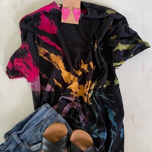 May include: A black tie-dye t-shirt with a colourful splatter pattern, paired with blue jeans and black sandals.