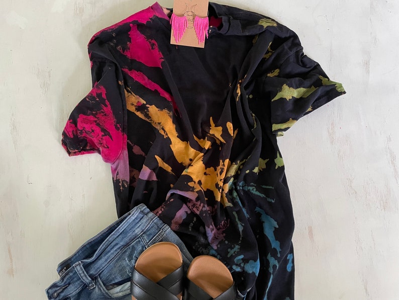 May include: A black tie-dye t-shirt with a colourful splatter pattern, paired with blue jeans and black sandals.