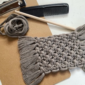 May include: A close-up of a gray macrame wall hanging with fringe on the bottom. The wall hanging is on a brown clipboard with a comb and a wooden dowel.