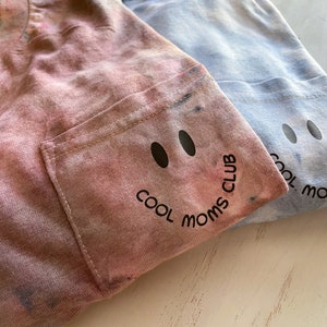 May include: Two tie-dye shirts with a pocket on the left side. The shirts have a black smiley face with the text "COOL MOMS CLUB" printed on the pocket.