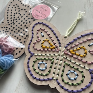 May include: A wooden butterfly shaped craft kit with colorful embroidery floss. The butterfly is partially stitched with purple, pink, yellow, green, and blue thread. The kit includes a wooden butterfly with pre-punched holes and embroidery floss.