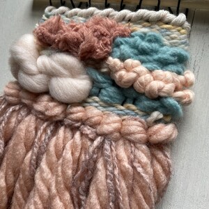 May include: A handcrafted woven wall hanging featuring a blend of textures and colors. The piece incorporates soft, fluffy yarn in shades of blush pink, cream, and teal, creating a tactile and visually appealing artwork. It is mounted on a natural wooden dowel.
