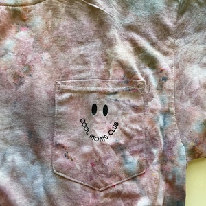 May include: A tie-dye t-shirt with a pocket featuring a black smiley face with the text "Cool Moms Club" on a light blue background.