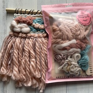 May include: A woven wall hanging with a light pink, blue, and white color scheme. The wall hanging is made of various textures of yarn and is mounted on a wooden dowel. A pink package next to it contains a Woven Wall Hanging Kit.