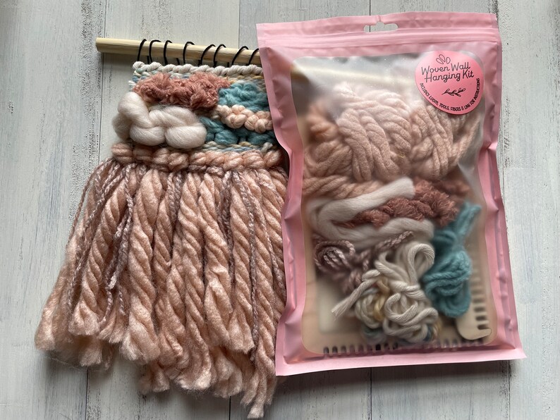 May include: A woven wall hanging with a light pink, blue, and white color scheme. The wall hanging is made of various textures of yarn and is mounted on a wooden dowel. A pink package next to it contains a Woven Wall Hanging Kit.