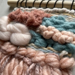 May include: Close-up of a woven wall hanging with a wooden dowel at the top. The textile features a mix of textures and colors, including white, teal, peach, and pink yarns. The design incorporates various weaving techniques, creating a tactile and visually appealing piece.
