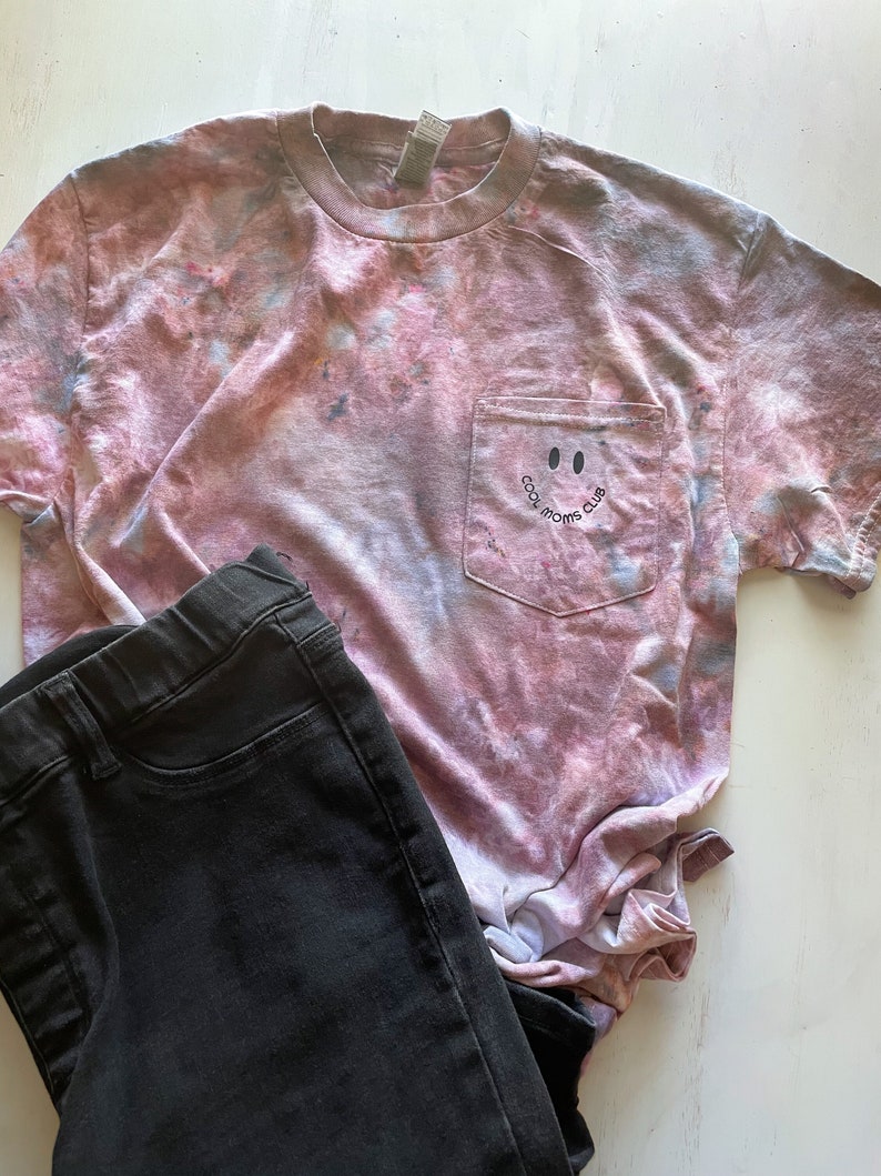 May include: A pink and grey tie-dye t-shirt with a pocket on the front. The pocket has a black smiley face and the text "Cool Moms Club".